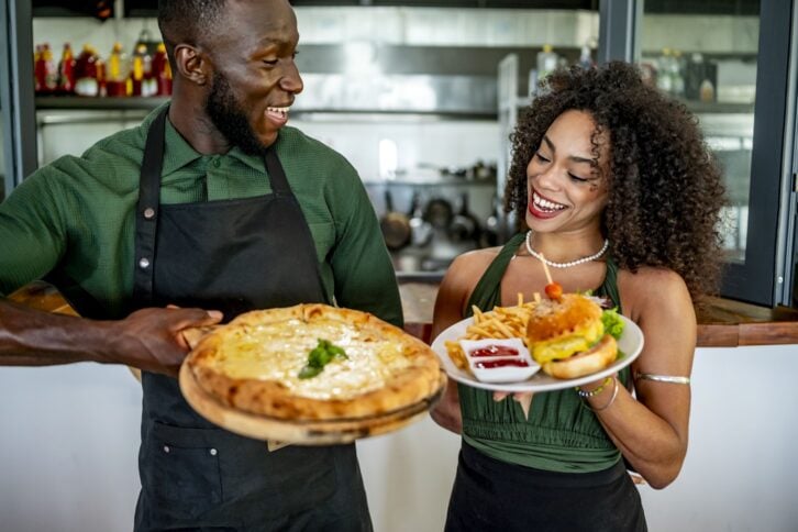 Smiling chefs holding delicious pizza and burger with fries in a restaurant kitchen, showcasing culinary skills and teamwork while enjoying their work environment.