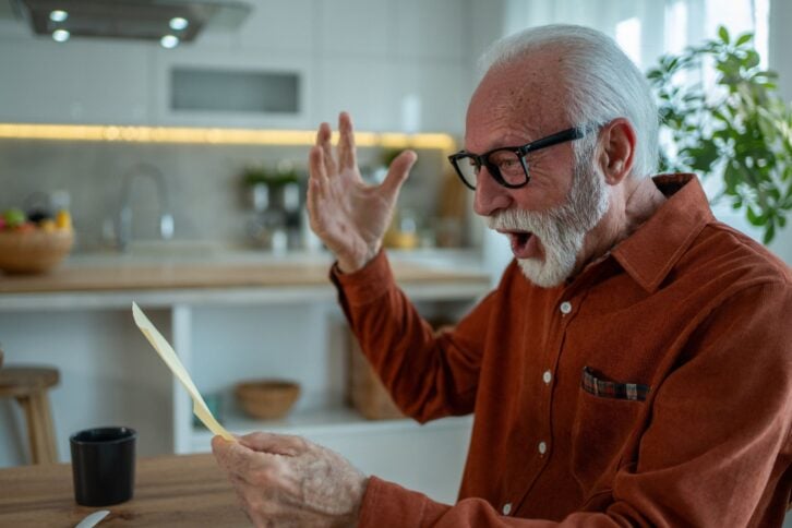 Surprised senior man reading a letter, reacting with astonishment and disbelief while sitting at a table in his modern kitchen, experiencing unexpected news that stirs deep emotions