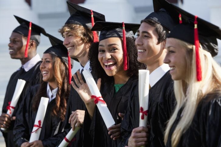 Multi-ethnic friends graduating together, in cap and gown.   Main focus on African American girl in middle, waving at camera.