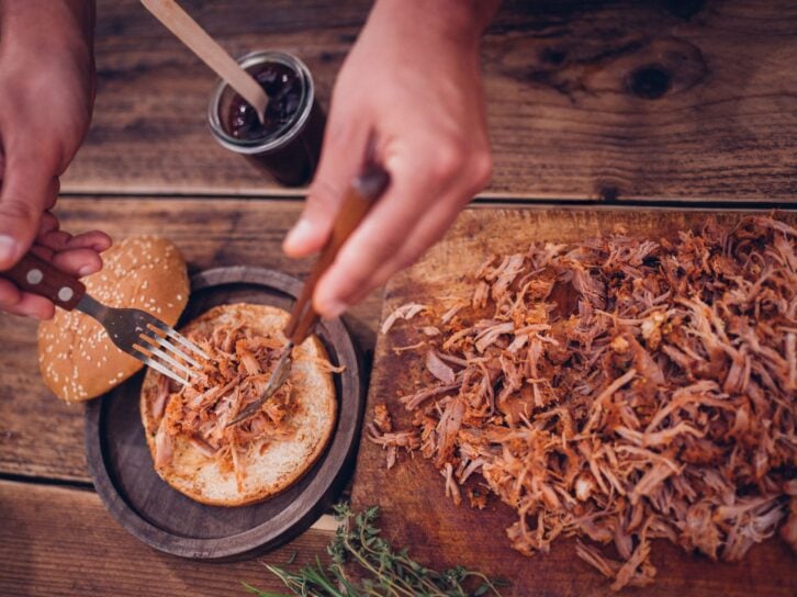 Overhead shot of some delicious pulled pork being placed on a toasted burger bun on a vontage wooden table with a jar of sauce nearby