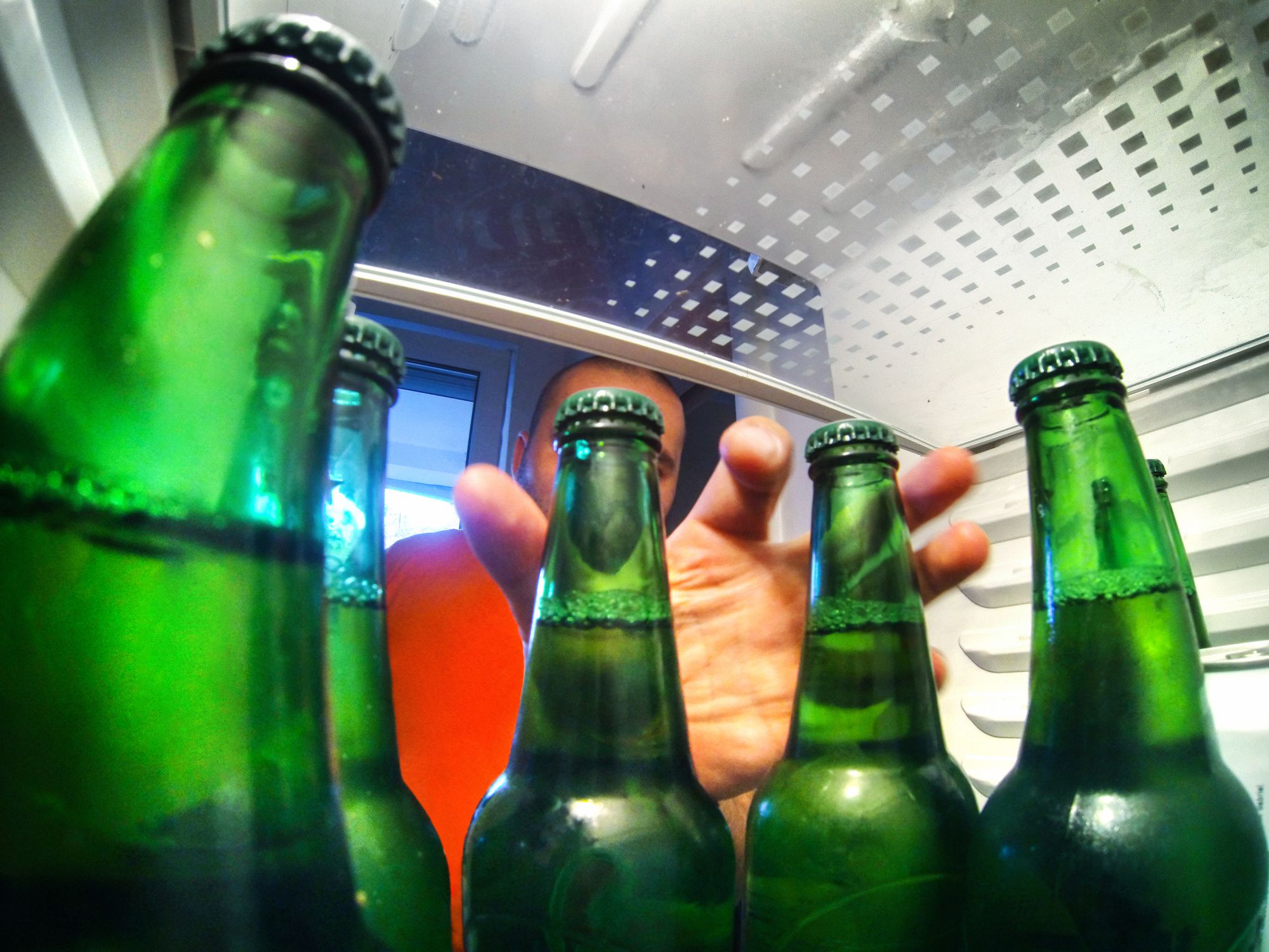 Closeup fisheye view of unrecognizable adult man grabbing couple of bottle of beer fromt his fridge. Shot from inside the fridge. Beer bottle are green and the man is wearing orange shirt.