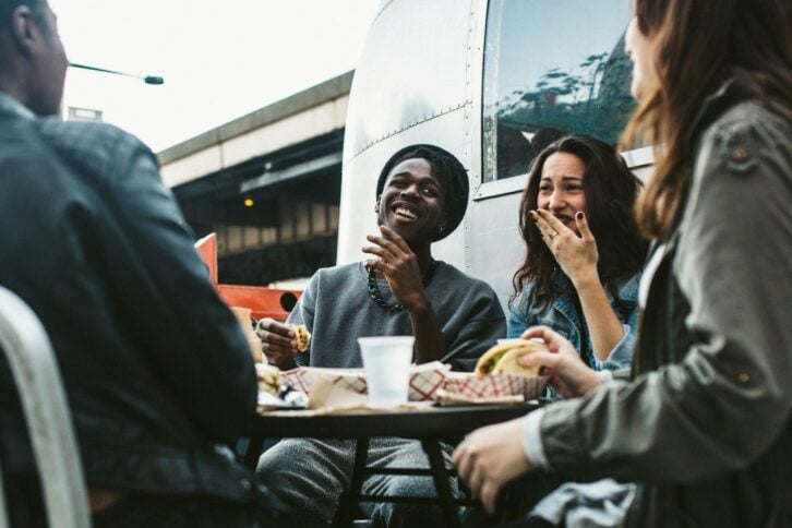 A multi-ethnic group of friends enjoy tacos and chips at a Mexican food cart in downtown New York City, New York.  They laugh and talk while eating their food, having fun sharing life and culture.  Horizontal image.