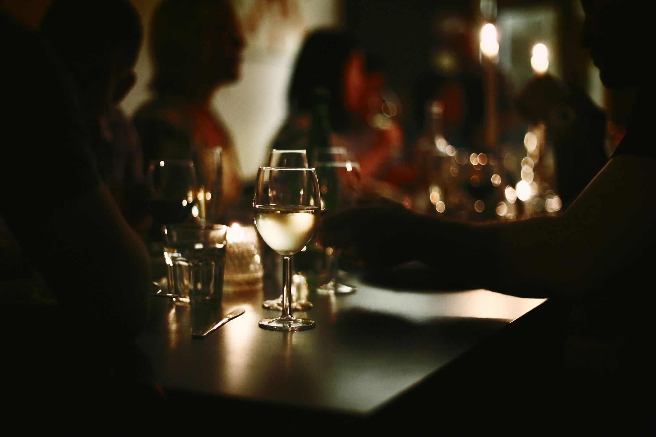 A darkened bistro with people socializing. Focus on a glass of chardonnay.