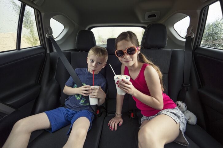 Two kids sitting in the back seat of their car eating an ice cream treat milkshake. Smiling and enjoying a sweet treat while sitting in their family automobile