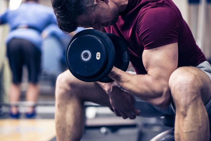 Muscular young man doing exercises for biceps muscles with dumbbells on in a gym.
