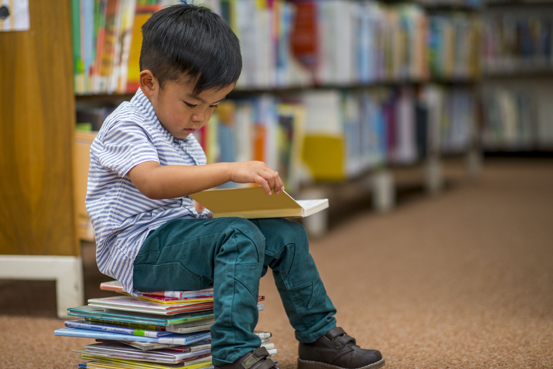 A little boy is sitting on a stack of books at the library and is reading.