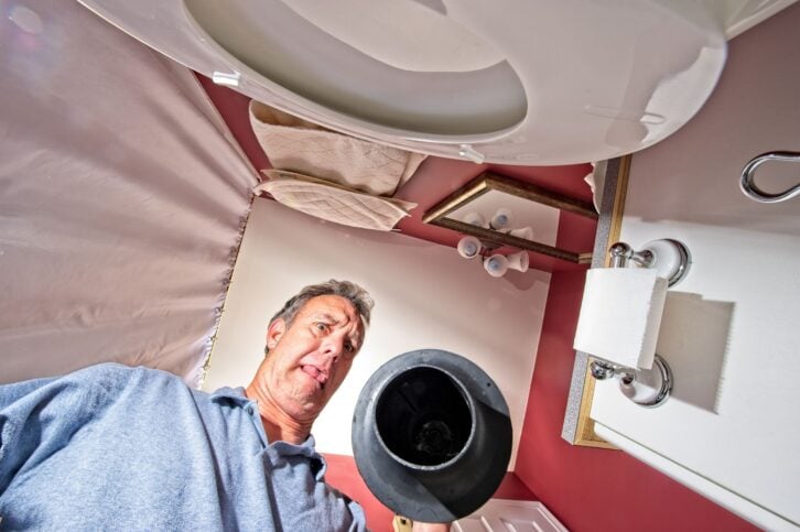 Unhappy senior man cleaning a toilet with a plunger.  View is upward from inside the toilet bowl and the man is making a face in disgust.