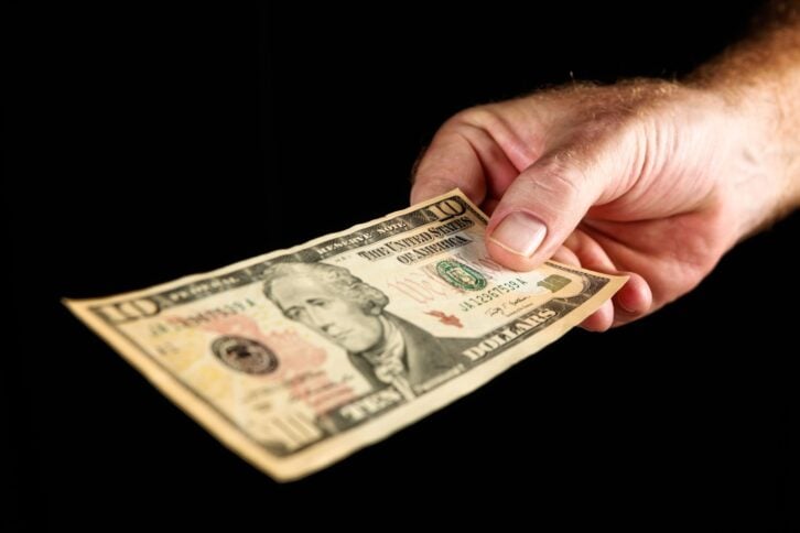 A man's hand holds out an American Ten Dollar bill against a black background with some copy space.