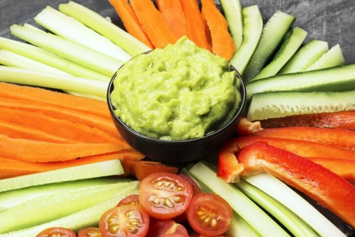 Traditional Latin American sauce guacamole in a bowl and various vegetables (carrots, tomatoes, cucumbers, celery) on a dark background.
