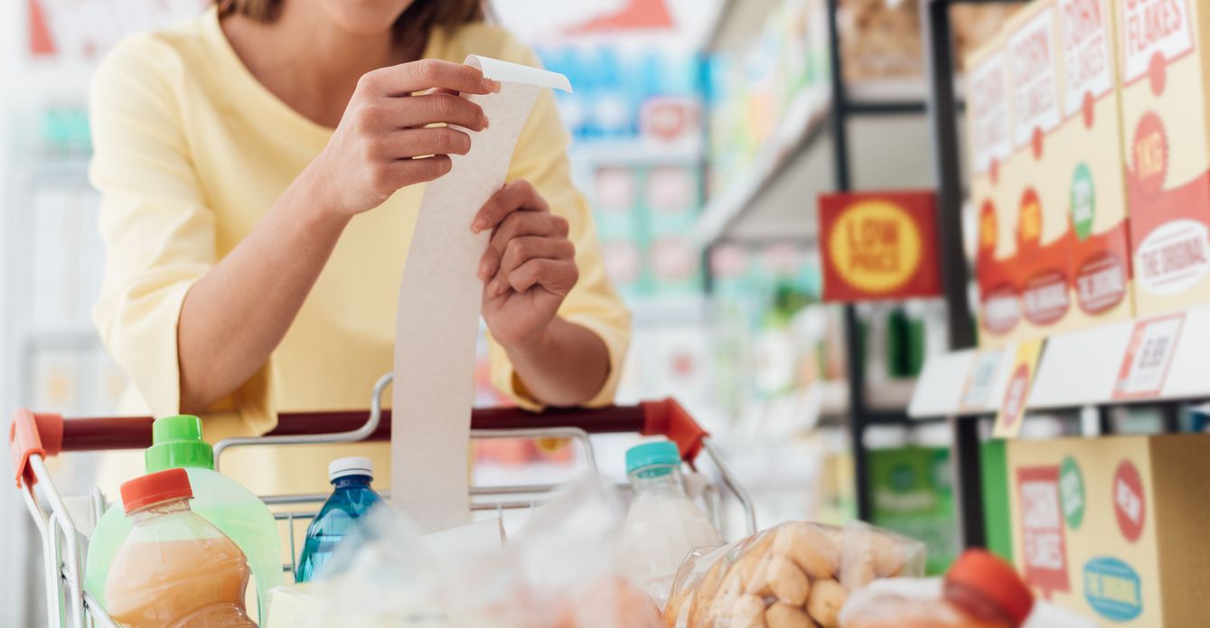 Smiling woman doing grocery shopping at the supermarket and checking a long receipt, she is leaning on the full cart