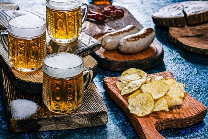 Three crystal mugs of golden beer with grilled bratwurst and homemade potato chips. Spotty blue background. Handmade wooden dishware. Close-up
