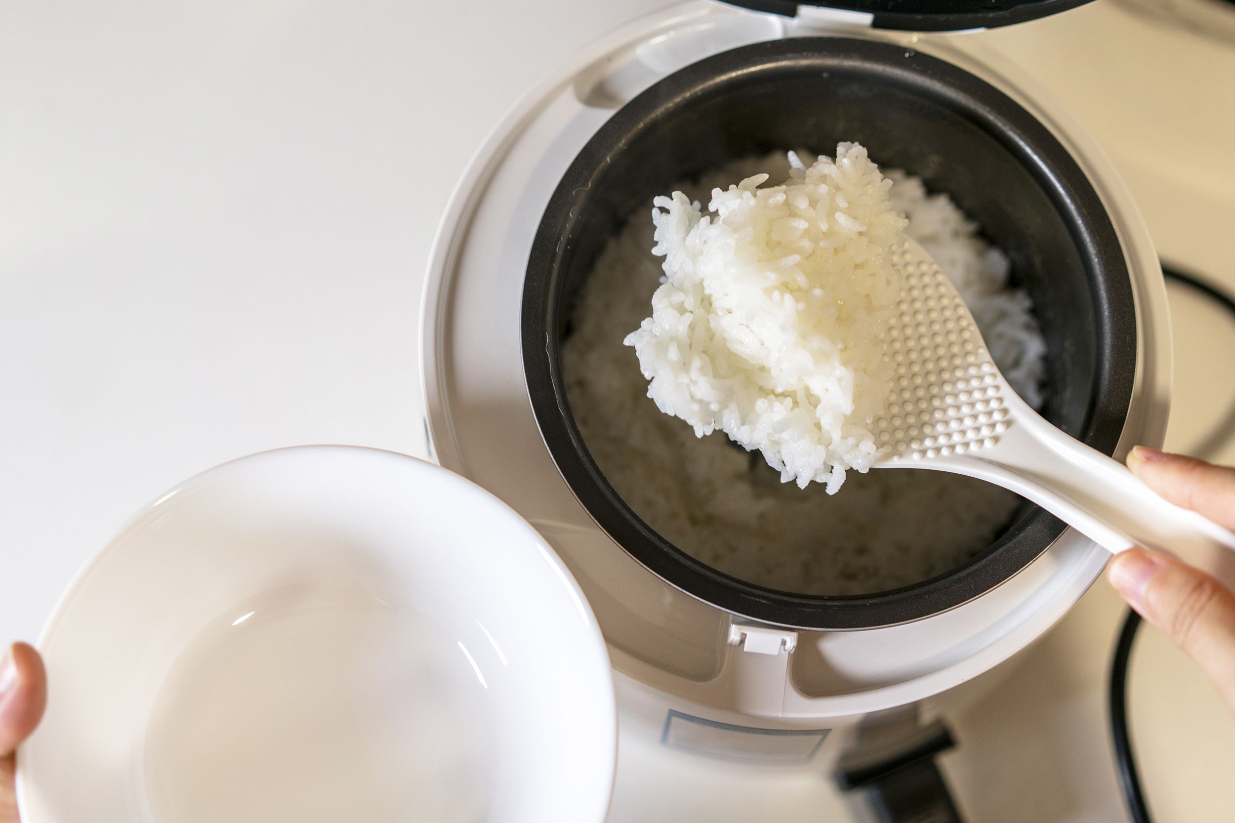 A person uses a white rice paddle to scoop cooked white rice from a rice cooker into a white bowl. The rice cooker lid is open and the rice appears fluffy and freshly made.