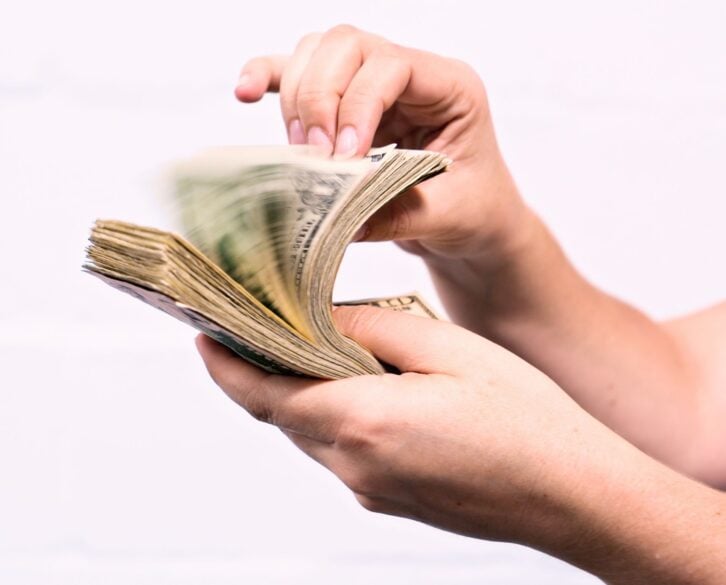 A woman's hand riffles through a thick bundle of American banknotes, creating motion blur.