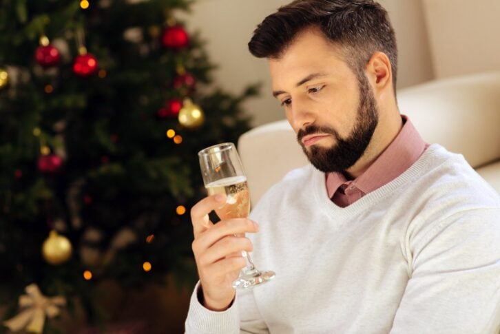 man looking uneasily at glass of champagne with christmas tree in background