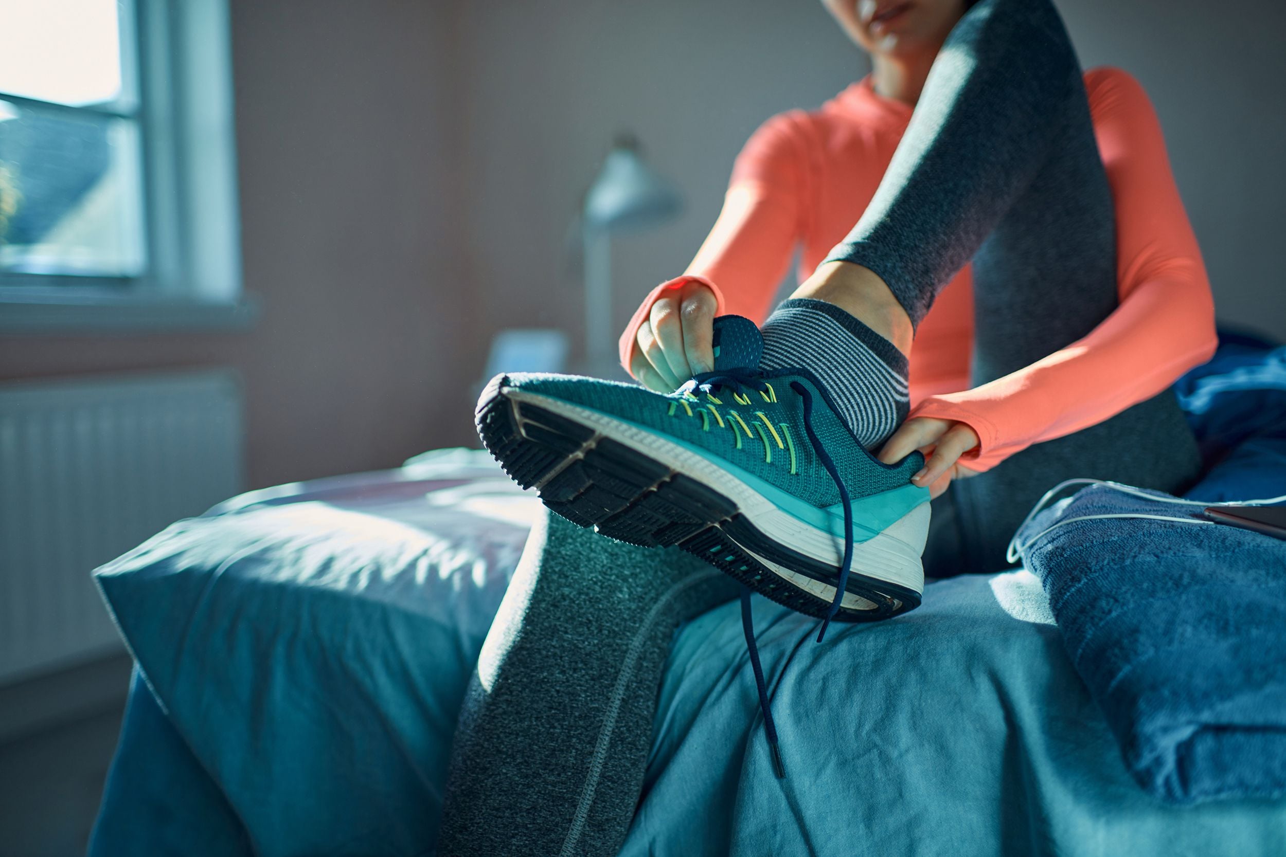 A person in athletic wear sits on a bed and ties the laces on one of their running shoes. Sunlight streams in through a window, and the room appears calm and tidy.