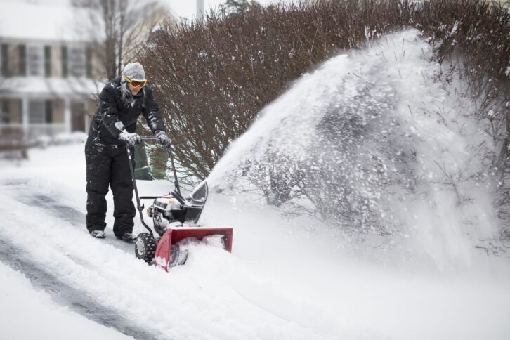 A man all bundled up pushing a snowblower.