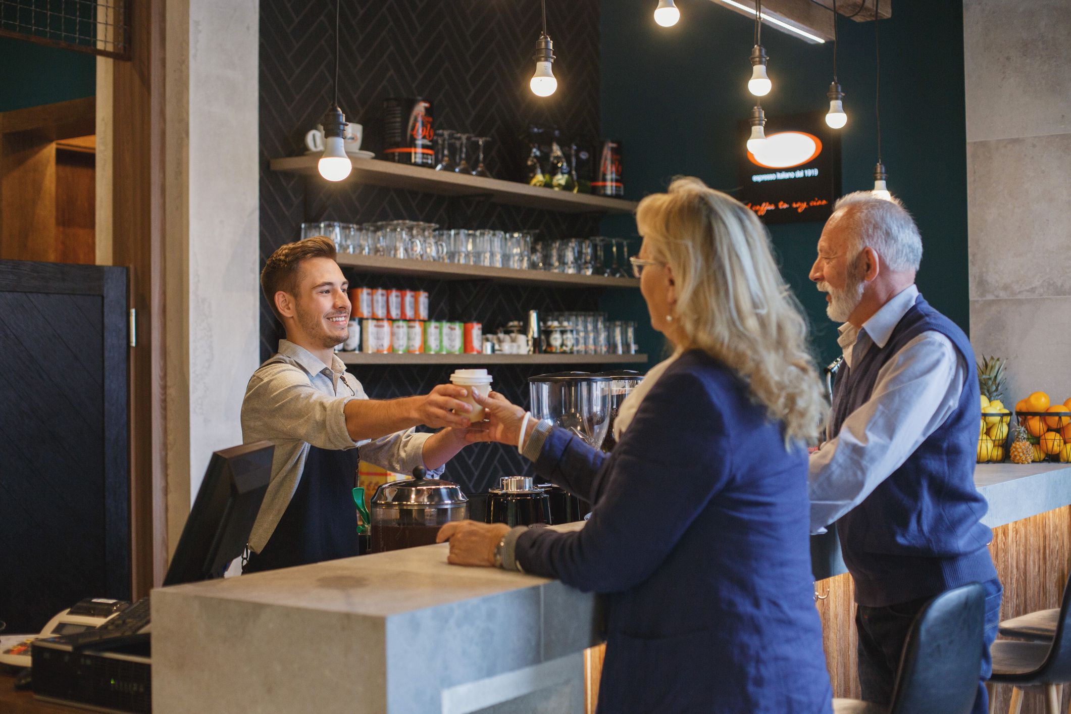 Smiling young barista is serving coffee to go to a senior couple.