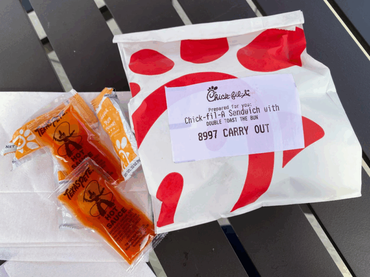 A Chick-Fil-A sandwich in a paper bag with hot sauce and honey packets