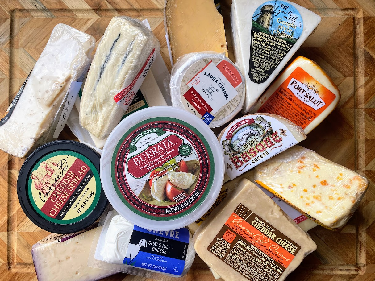 An assortment of various cheeses from Trader Joe's, both sliced and whole, displayed on a wooden cutting board. Labels show different types, including burrata, cheddar, Port Salut, brie, goat cheese, and blue cheese.