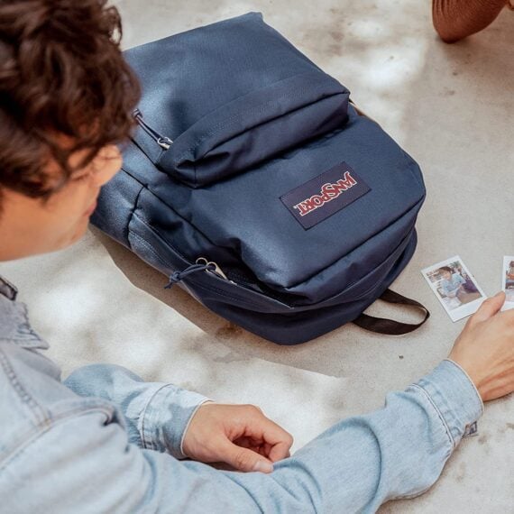 A jansport backpack laying on the ground while a young man in a denim jacket looks at photos.