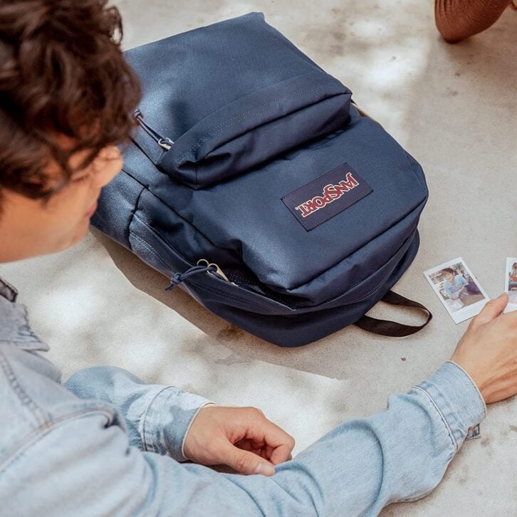 A jansport backpack laying on the ground while a young man in a denim jacket looks at photos.