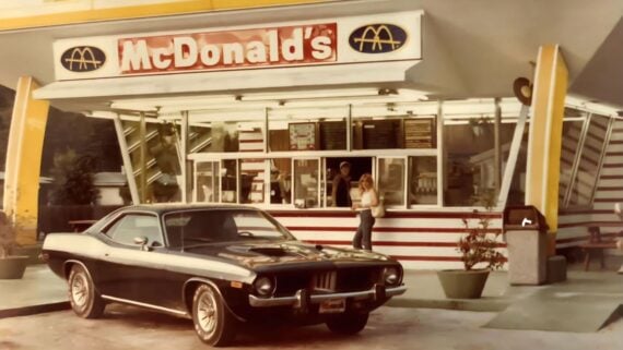 A vintage black muscle car parked in front of a McDonald’s restaurant with a person waiting at the walk-up service window.