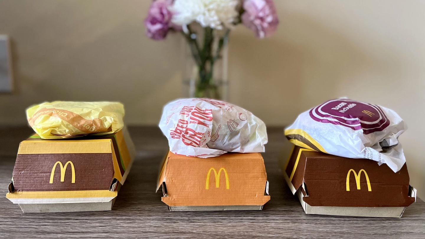 Three McDonald’s burgers in wrappers and boxes are arranged on a wooden table, with a vase of white and pink flowers in the blurred background.