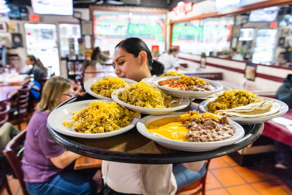 A server carries a large tray filled with plates of Mexican food, including rice, beans, and tortillas, in a busy restaurant with diners seated at tables in the background.