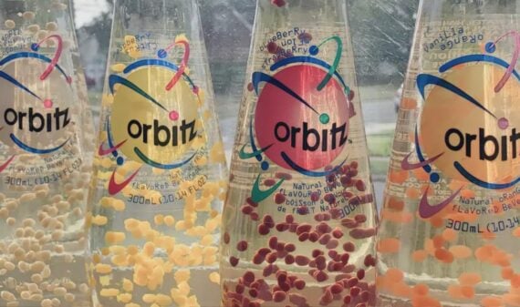 Four bottles of Orbitz soda with floating colored gel balls, each with a colorful label and scientific-themed design, arranged in a row on a window sill with a blurred outdoor background.
