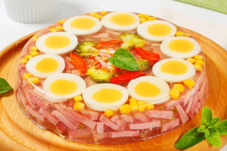 A round savory aspic on a wooden plate, topped with slices of boiled eggs, corn, broccoli, and red bell pepper, with assorted meats and vegetables set in clear gelatin. Green herbs decorate the plate.