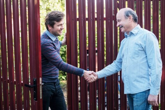 A younger man and an older man shake hands through a maroon metal gate.