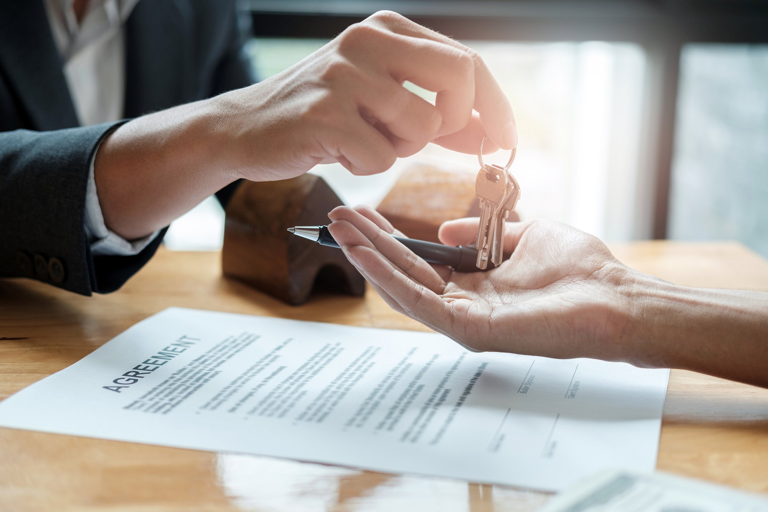 A person hands over a set of keys to another person above a signed agreement document on a wooden table, symbolizing a transaction or property transfer.