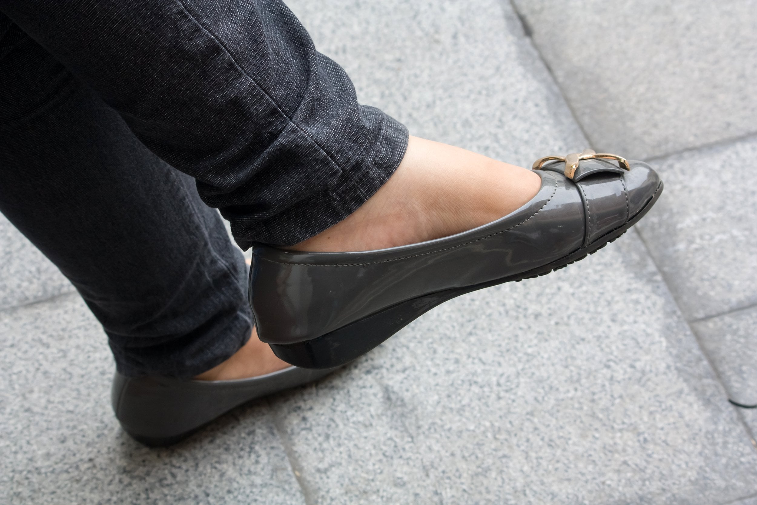 A person wearing gray shiny flats with gold accents and black jeans stands on light gray pavement, with one foot slightly raised and crossed over the other.