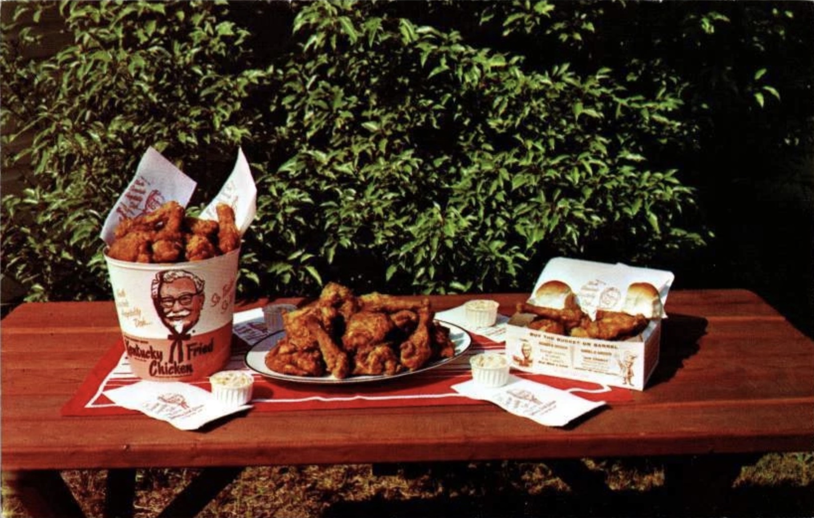 A picnic table outdoors holds a large KFC bucket of fried chicken, a plate of fried chicken pieces, a box with KFC sandwiches, sauce cups, napkins, and a placemat, with green bushes in the background.