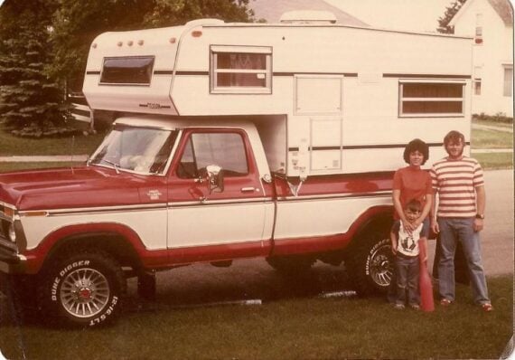 A family of three stands by a red and white pickup truck with a camper attached, parked on a suburban street with houses and trees in the background.