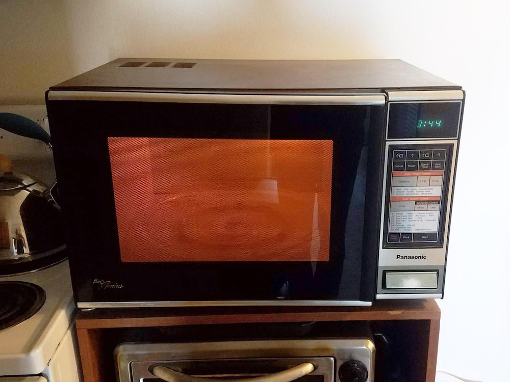 A Panasonic microwave oven with an orange-lit interior sits on a wooden shelf above a toaster oven. The digital display shows the time 3:44, and various buttons are visible on the control panel.