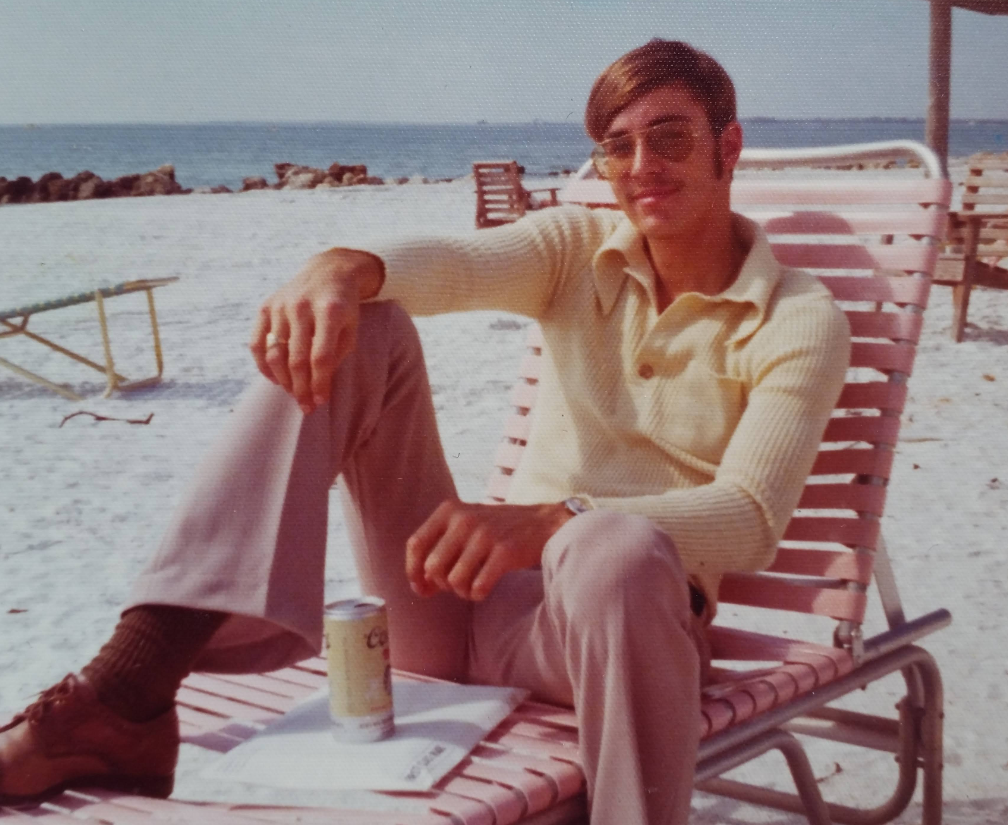 My dad in the 70s wearing a sweater, pants and leather shoes on the beach drinking a Coors