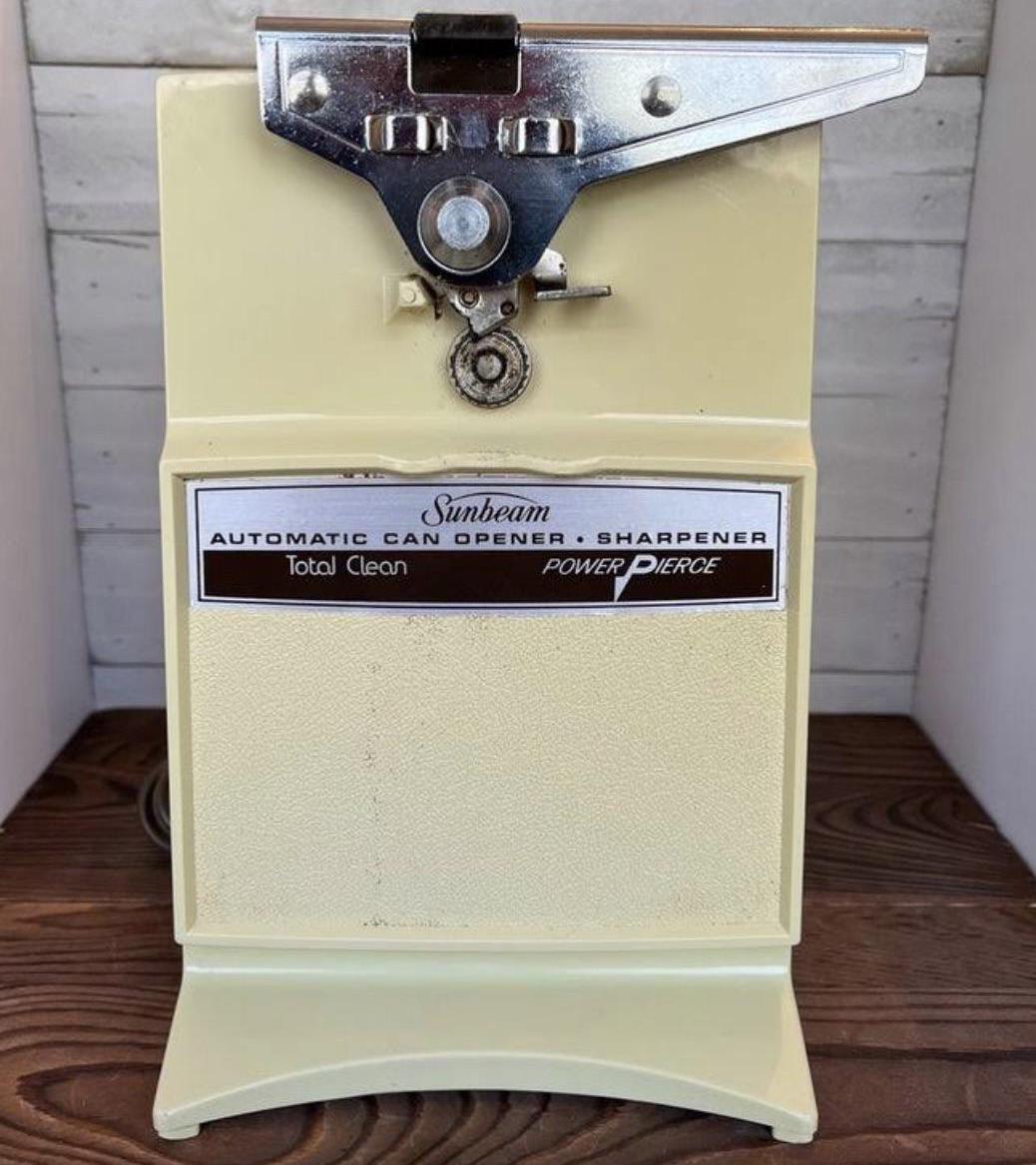 A vintage beige Sunbeam Power Pierce automatic can opener and sharpener with a metal lever and label on the front, sitting on a wooden surface against a light background.