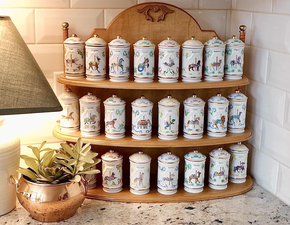 A wooden spice rack holds three rows of matching decorative ceramic jars, each with painted animal designs. A copper pot with a green plant sits to the left, under a lamp, on a granite countertop with white tiled walls.