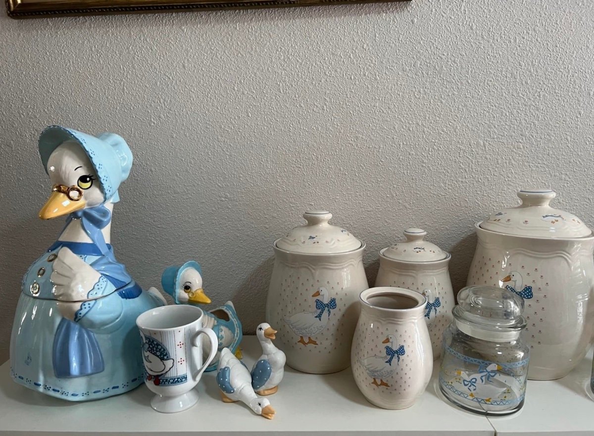A collection of ceramic kitchenware decorated with blue-and-white goose designs, including cookie jars, a pitcher, a mug, two figurines, and a glass jar, all arranged on a white shelf against a plain wall.