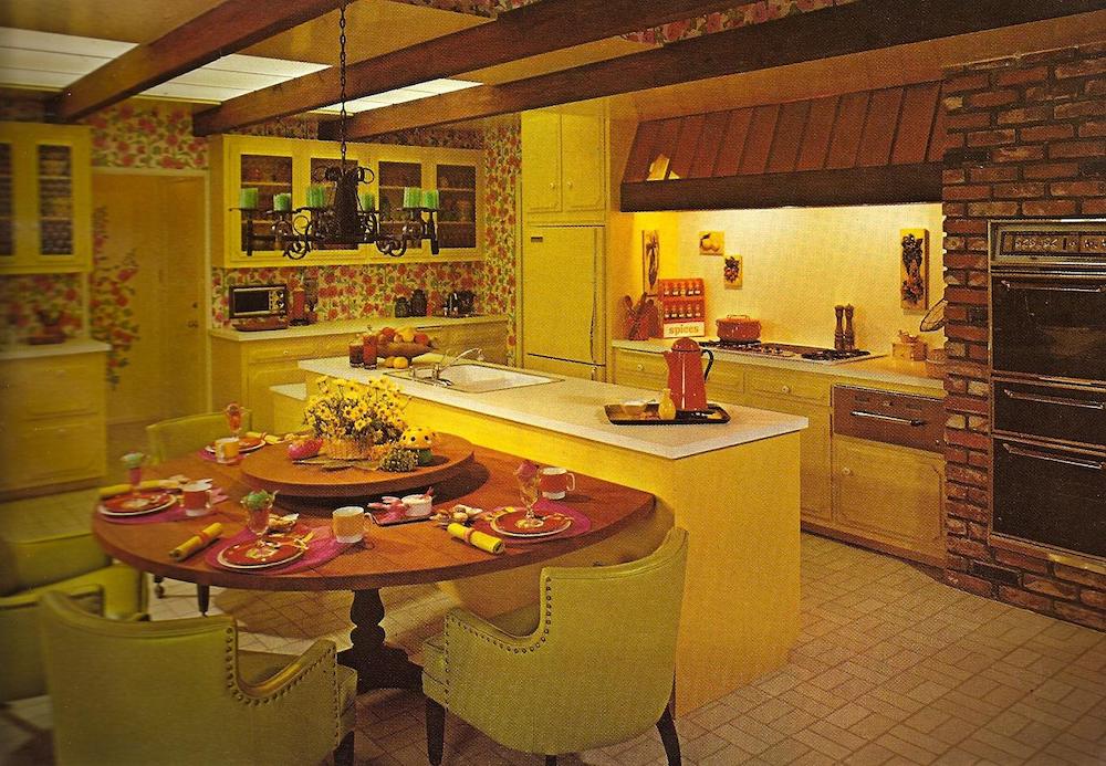 A vintage kitchen with yellow cabinets, floral wallpaper, a brick oven wall, and exposed beams. A round wooden table with green chairs is set for a meal beside a white kitchen island. The room has a cozy, retro feel.