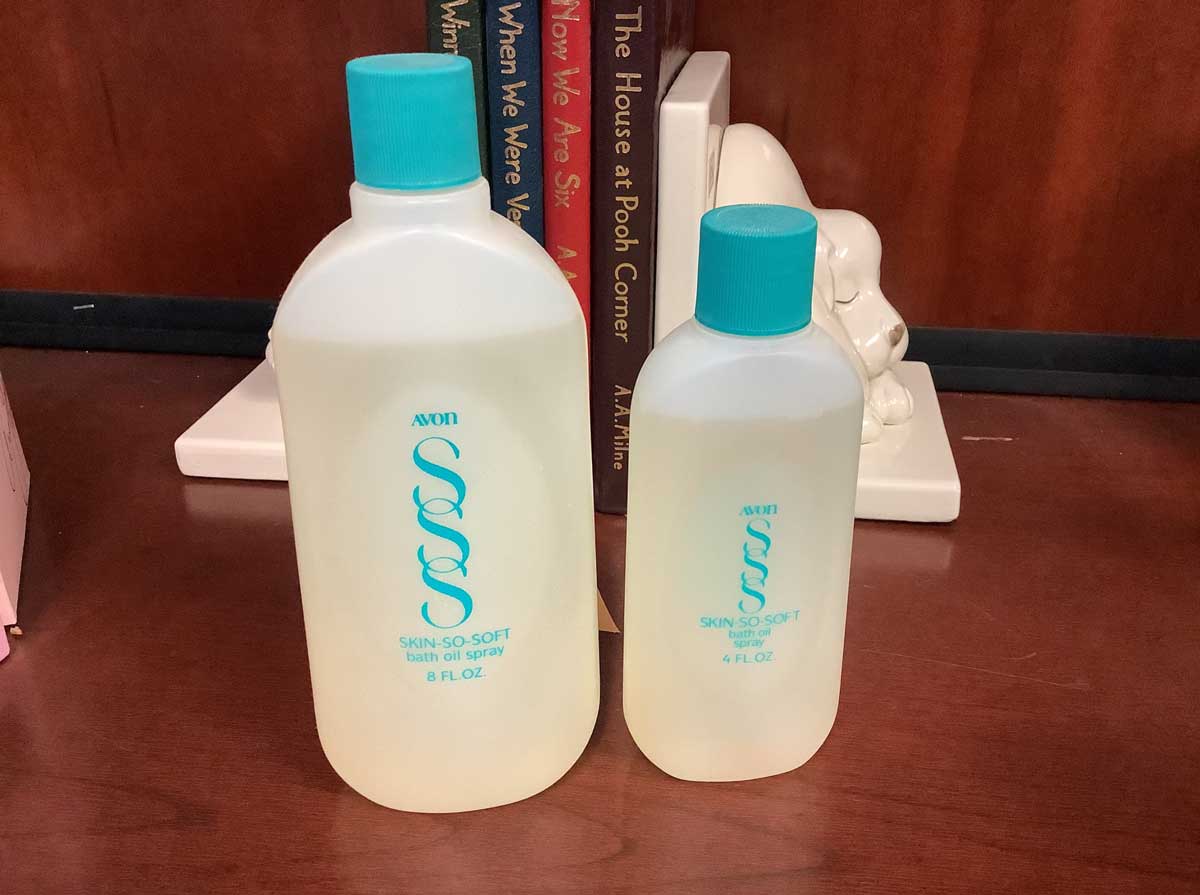 Two bottles of Avon Skin-So-Soft bath oil, one 8 fl. oz. and one 4 fl. oz., evoke a touch of the 1980s as they sit on a wooden surface in front of several upright books.