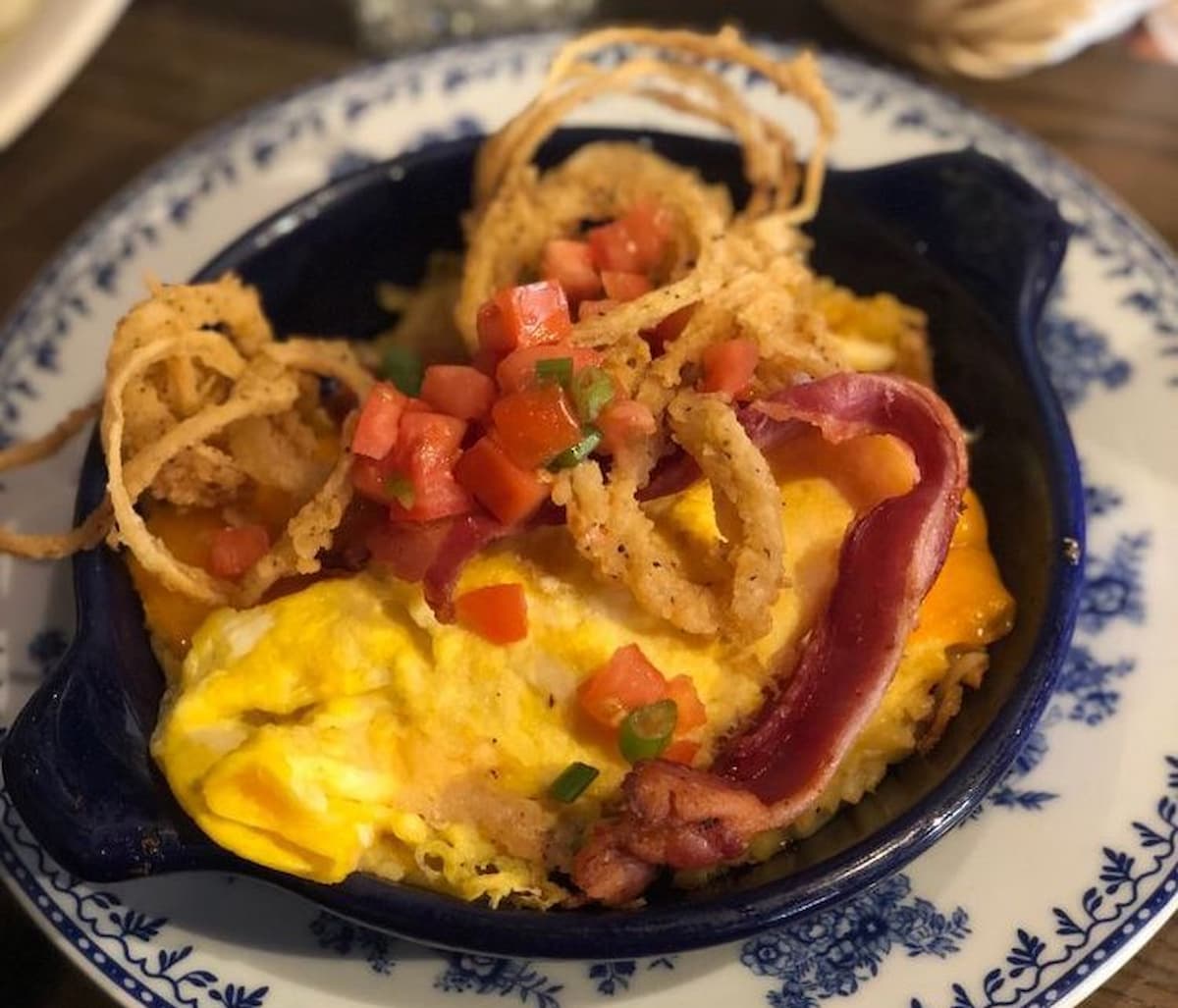 A blue dish inspired by Cracker Barrel with scrambled eggs, crispy fried onion strings, diced tomatoes, chopped green onions, melted cheese, and bacon pieces on a white plate with a blue floral pattern.