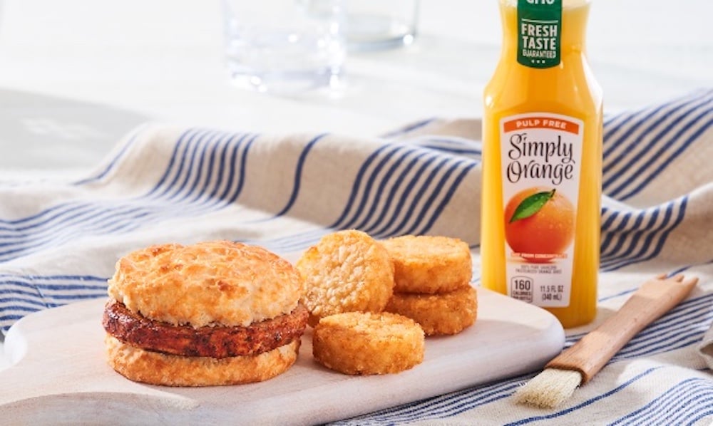 A new fast-food breakfast sandwich with a biscuit and sausage patty, four hash brown rounds, and a bottle of Simply Orange juice are arranged on a wooden board, sitting on a striped cloth.