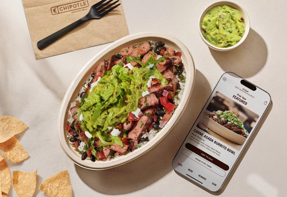 A Chipotle burrito bowl with steak, black beans, salsa, and guacamole sits next to a cup of guacamole, tortilla chips, a napkin with a fork, and a smartphone displaying a new fast-food Chipotle order screen.