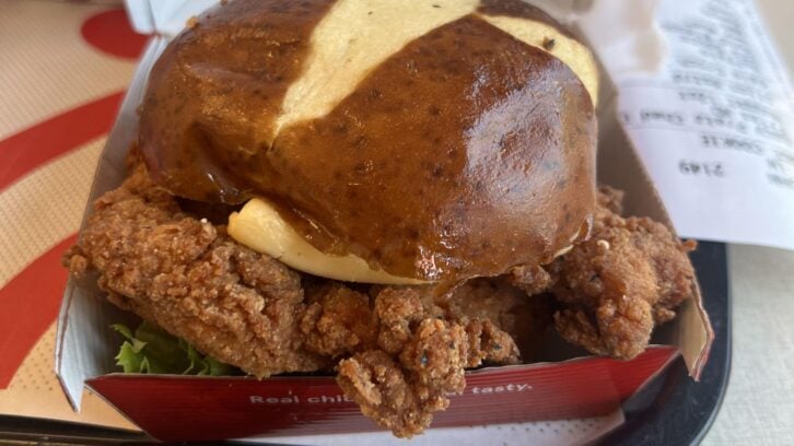 A Chick-fil-A crispy fried chicken sandwich with lettuce on a pretzel bun, served in a red box with a receipt and tray in the background—a must-try from the new fast-food menu.