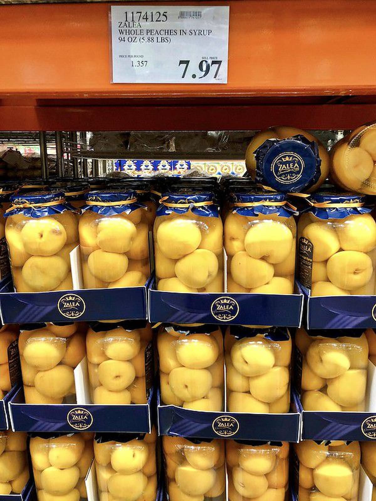 Large jars of whole peaches in syrup, each with a blue lid and label, are stacked on store shelves. The price sign above reads $7.97 for a 94 oz jar—a sweet find for those looking for Costco shopping tips.