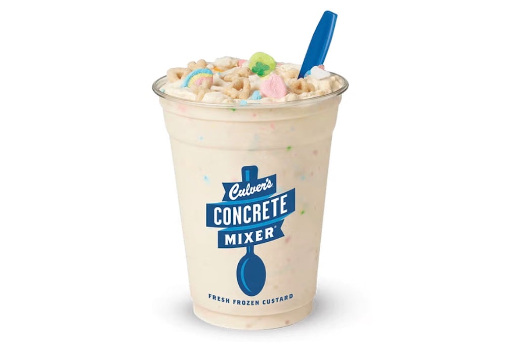 A clear cup of Culver’s new fast-food Concrete Mixer frozen custard, topped with colorful marshmallow cereal pieces and a blue spoon. The cup features the signature blue Culver's Concrete Mixer logo.