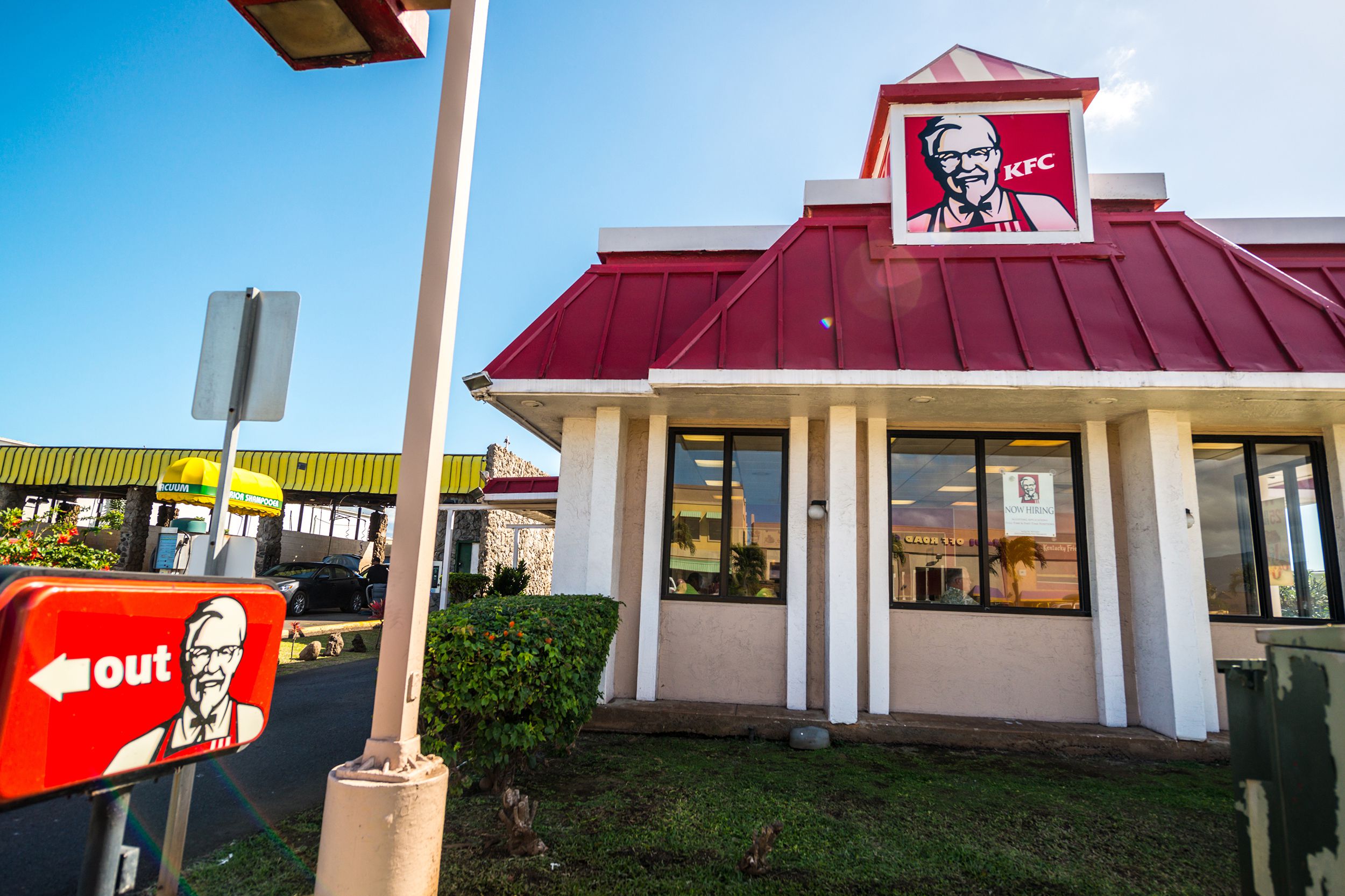Exterior of KFC fast food restaurant, Maui, Hawaii, garden and 'out' sign for the drivethru with another establishment in the background, against a bright blue sky