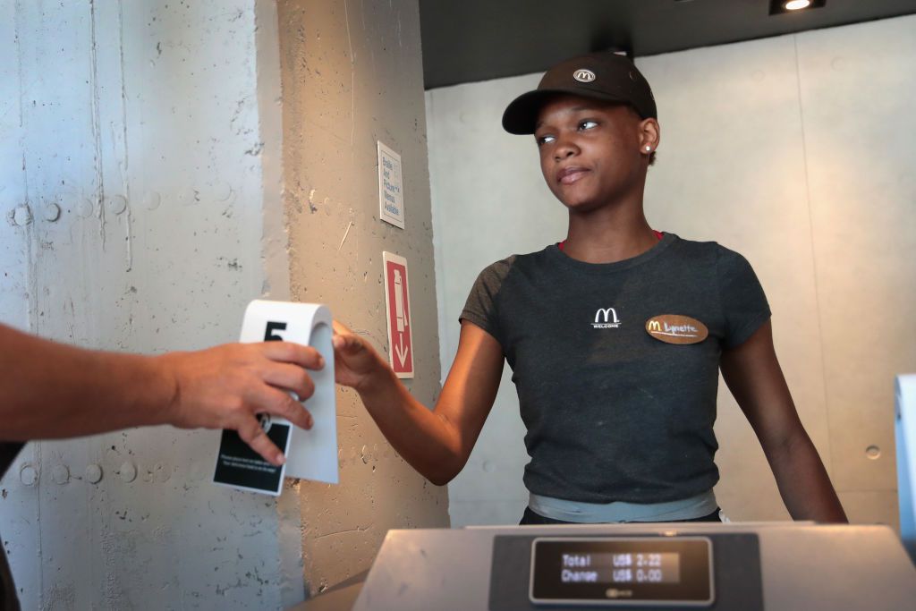 A McDonald's employee in uniform hands a receipt to a customer across the counter. The employee stands near a concrete wall, looking focused, and there is a digital register in the foreground.
